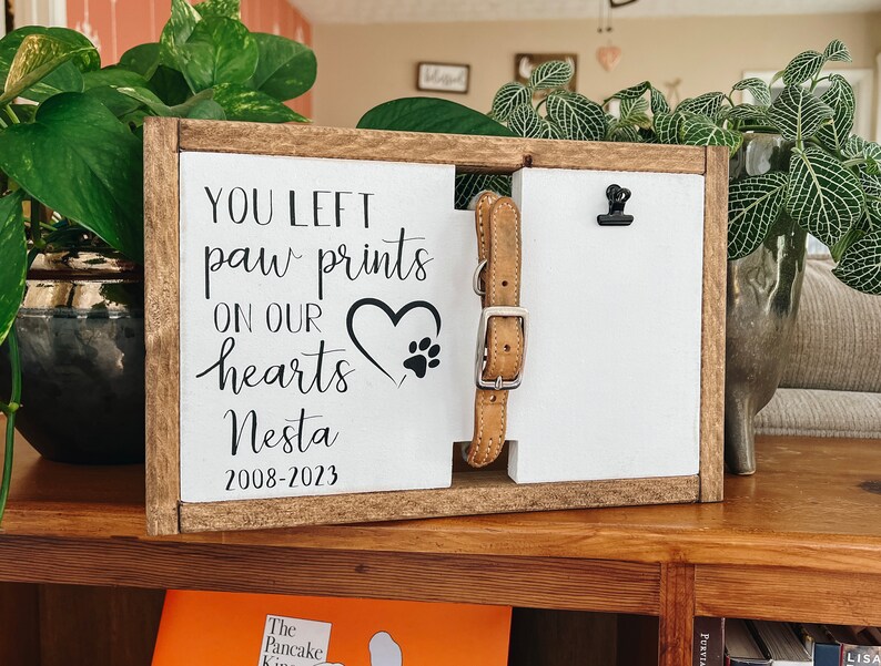 May include: A wooden sign with the text "You left paw prints on our hearts Nesta 2008-2023". A heart with paw prints is in the center of the sign. A brown leather dog collar is attached to the sign with a black clip.