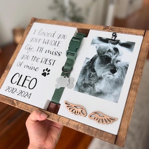 May include: A wooden frame with a white background and a photo of a small dog. The frame has a green dog collar with a silver tag attached. The text "I loved you your WHOLE life, I'll miss you the REST of mine CLEO 2012-2024" is printed on the frame. Two wooden angel wings are attached to the bottom of the frame.