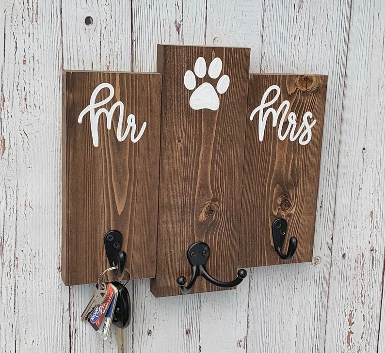Op de afbeelding: Een houten sleutelhanger met twee haken en de woorden "Mr." en "Mrs." aan weerszijden van een pootprint.