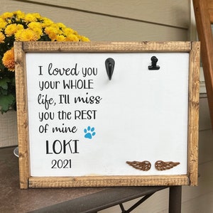 May include: A white wooden sign with a brown frame, featuring the text "I loved you your WHOLE life, I'll miss you the REST of mine LOKI 2021". A blue paw print and two brown wooden wings are also visible on the sign.