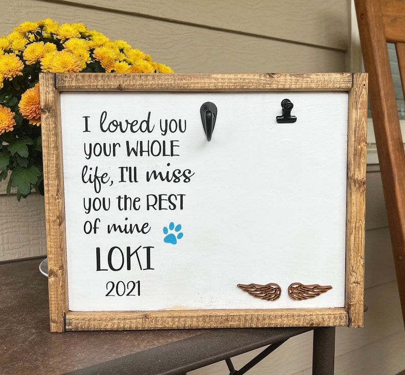 May include: A white wooden sign with a brown frame, featuring the text "I loved you your WHOLE life, I'll miss you the REST of mine LOKI 2021". A blue paw print and two brown wooden wings are also visible on the sign.