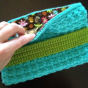 May include: A turquoise crocheted clutch with a green crocheted stripe and a brown fabric lining with a floral print. The clutch has a magnetic closure.
