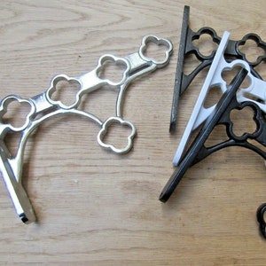 May include: A selection of decorative metal shelf brackets in different colours. The brackets have a clover-like design and are shown in silver, black, and white. The brackets are designed to be mounted on a wall to support shelves.