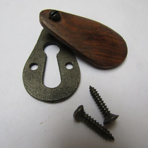 May include: A keyhole cover with a wooden escutcheon. The cover is made of dark wood and has a rounded shape. The keyhole cover is attached to a metal plate with two screws.