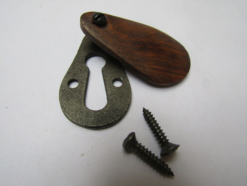 May include: A keyhole cover with a wooden escutcheon. The cover is made of dark wood and has a rounded shape. The keyhole cover is attached to a metal plate with two screws.