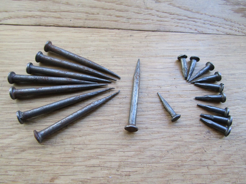May include: A collection of 18 antique iron nails with pointed tips and round heads. The nails are arranged in two groups on a wooden surface.