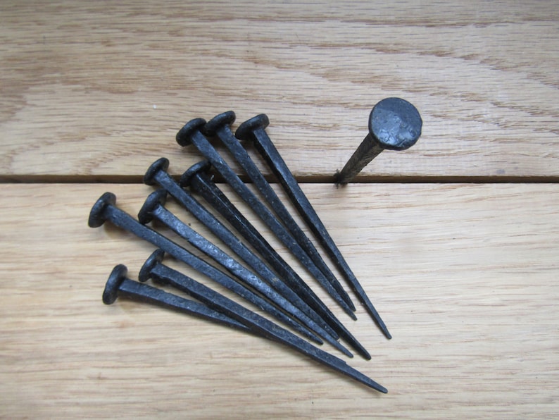 May include: A set of ten black wrought iron nails with rounded heads. The nails are arranged in a fan shape on a wooden surface.