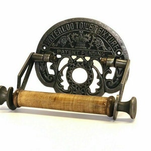 May include: Cast iron toilet paper holder with a wooden roller and a decorative design. The holder has the text "Waterloo Toilet Fixture Patent Pending" on the front.