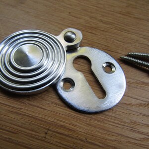 May include: A silver keyhole cover with a round, concentric circle design. The cover is attached to a smaller, oval-shaped keyhole plate. Two silver screws are included.