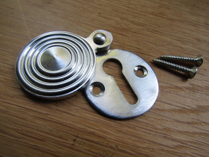 May include: A silver keyhole cover with a round, concentric circle design. The cover is attached to a smaller, oval-shaped keyhole plate. Two silver screws are included.