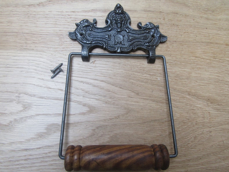 May include: A cast iron toilet paper holder with a decorative scroll design and a wooden roller. The holder is mounted on a wall with two screws.