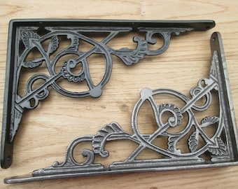 Puede incluir: Dos soportes de estante de hierro fundido negro con un diseño floral ornamentado. Los soportes tienen forma de ángulo recto y presentan un patrón de volutas decorativas.