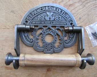 May include: Cast iron toilet roll holder with a decorative design and the text "St Pancras Fixture 18 x 83 WBF & S/E LONDON". The holder has a wooden roller and is mounted on a wooden surface.