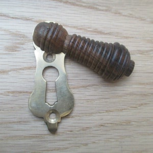 May include: A brass keyhole cover with a wooden knob. The knob is dark brown and has a ribbed texture. The keyhole cover is ornate and has a decorative design.