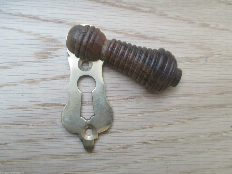 May include: A brass keyhole cover with a wooden knob. The knob is dark brown and has a ribbed texture. The keyhole cover is ornate and has a decorative design.