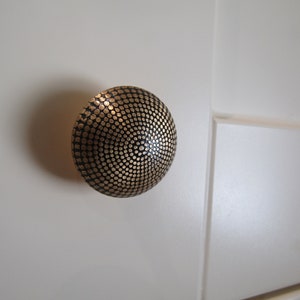 May include: A round cabinet knob featuring a dark metallic finish with a textured surface composed of numerous small, raised circular dots arranged in a concentric pattern radiating from the center point of the dome-shaped hardware piece.