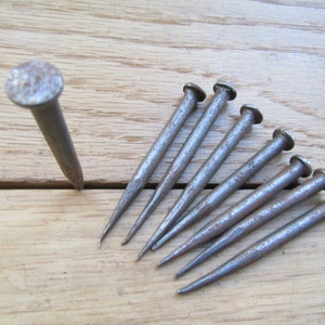 May include: A set of eight antique, rusty, iron nails with pointed tips. The nails are arranged in a fan shape on a wooden surface.