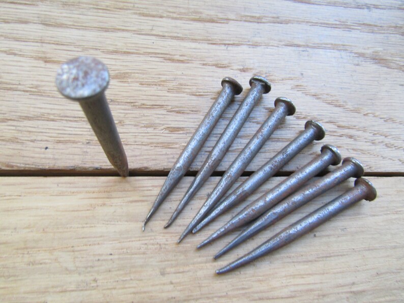 May include: A set of eight antique, rusty, iron nails with pointed tips. The nails are arranged in a fan shape on a wooden surface.