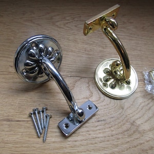 Op de afbeelding: Twee decoratieve metalen plankbeugels met een bloemmotief. Een beugel is verchroomd en de andere is gepolijst messing. Beide beugels worden geleverd met schroeven.