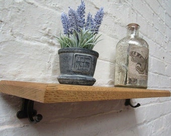 May include: A wooden shelf with black metal brackets is mounted on a white brick wall. A small potted plant with lavender-colored flowers and a decorative glass bottle with a butterfly design are displayed on the shelf.