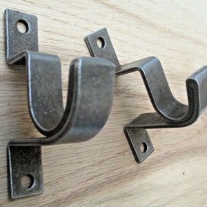 Könnte beinhalten: Zwei Gardinenstangenhalter aus Metall im antiken Stil. Jeder Halter ist dunkelgrau und verwittert, mit einer U-förmigen Krümmung und einer rechteckigen Montageplatte mit einem Loch für Schrauben. Die Halter sind für die Aufnahme einer Gardinenstange konzipiert.