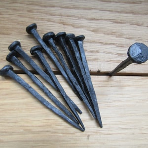 May include: A set of ten black wrought iron nails with rounded heads. The nails are arranged in a fan shape on a wooden surface.