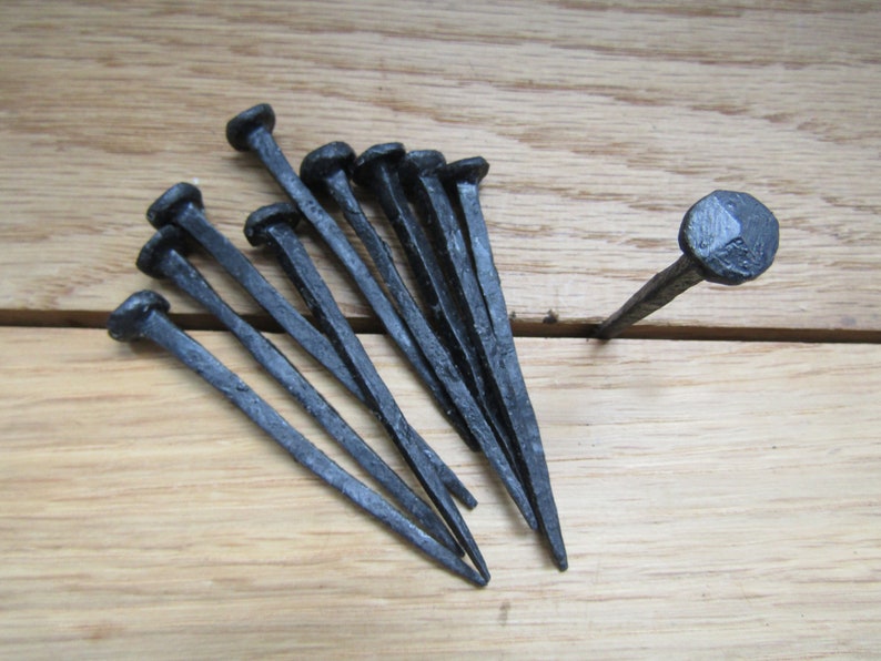 May include: A set of ten black wrought iron nails with rounded heads. The nails are arranged in a fan shape on a wooden surface.