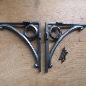 Puede incluir: Dos escuadras de estantería de hierro fundido con acabado gris. Cada escuadra tiene un diseño decorativo de volutas y un acento circular. Se incluyen varios tornillos oscuros. Las escuadras se muestran sobre una superficie de madera clara.