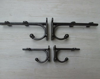 Puede incluir: Cuatro ganchos de pared de hierro fundido gris oscuro. Cada gancho tiene una sección inferior curvada con un extremo en forma de bola y una barra horizontal con detalles decorativos. Los ganchos están montados sobre una base cuadrada.