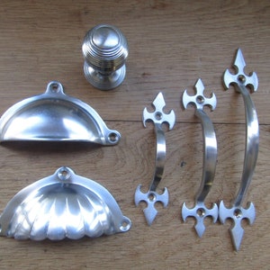 May include: A selection of silver-coloured cabinet hardware, including a round knob, two shell-shaped pulls, and three decorative handles with pointed ends. Several screws are also visible. The hardware is arranged on a wooden surface.