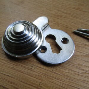 May include: Silver keyhole cover with a decorative, ridged, round escutcheon. The cover is designed to be mounted on a door or cabinet. Two screws are included.