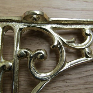 May include: A gold metal shelf bracket with a decorative, ornate design. The bracket has a scrolled, floral pattern and is made of cast metal.