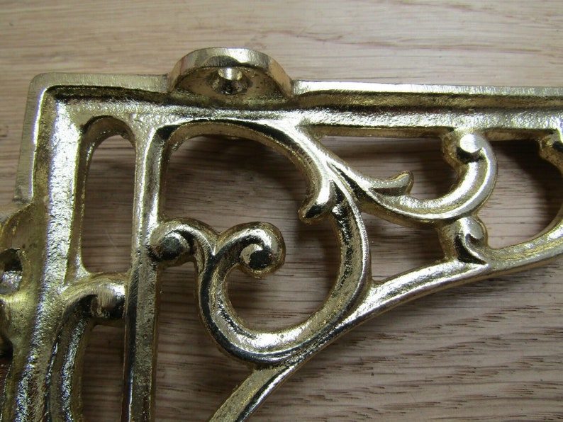 May include: A gold metal shelf bracket with a decorative, ornate design. The bracket has a scrolled, floral pattern and is made of cast metal.