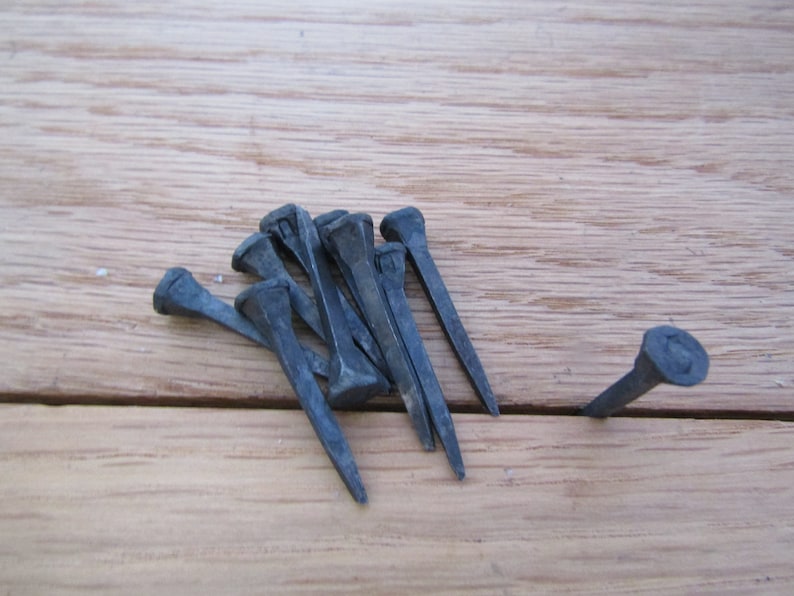 May include: A group of ten black metal nails with square heads, lying on a wooden surface. One nail is standing upright.
