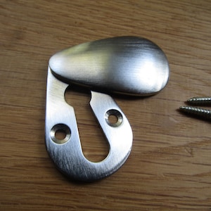 May include: A silver keyhole cover with a rounded, teardrop-shaped flap. The cover is designed to conceal a keyhole and is secured with two screws.