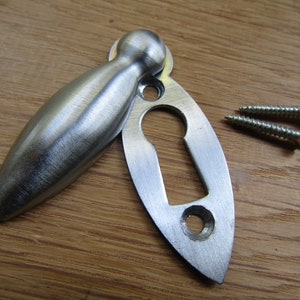 May include: A silver keyhole cover with a matching escutcheon. The cover is shaped like a teardrop and has a rounded top. The escutcheon is oval-shaped and has a keyhole cut out. Two screws are included.