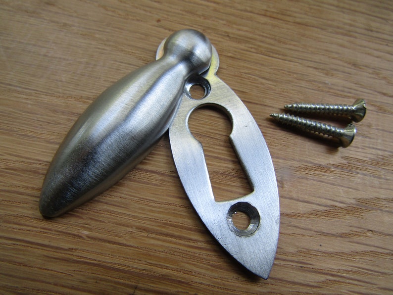 May include: A silver keyhole cover with a matching escutcheon. The cover is shaped like a teardrop and has a rounded top. The escutcheon is oval-shaped and has a keyhole cut out. Two screws are included.