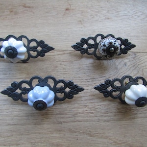 May include: Four decorative cabinet pulls with ornate, dark metal backplates. Two pulls have white, flower-shaped knobs, one has a light blue knob, and one has a patterned knob with brown and white details. The pulls are set against a light wood surface.