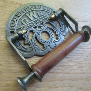 May include: Cast iron toilet paper holder with a wooden roller and a Great Western Railway logo. The holder is painted in a silver color and has a decorative design. The logo reads "Great Western Railway" and "GWR Patent Pending".