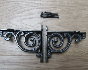 Puede incluir: Dos escuadras de estantería ornamentadas de hierro fundido gris oscuro con un diseño en espiral. Las escuadras en forma de L incluyen orificios de montaje. También hay un pequeño montón de tornillos, lo que sugiere un juego completo para la instalación.