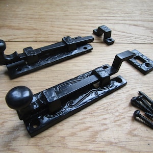 May include: Two black metal door latches with decorative details. Each latch has a round knob and a hook-shaped arm. The latches are shown on a wooden surface with screws.