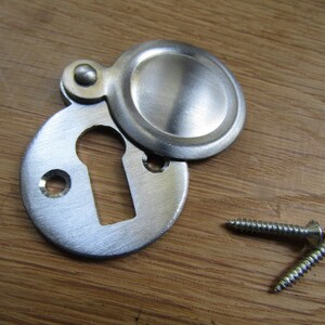 May include: A silver keyhole cover with a round, hinged lid. The cover is attached to a wooden surface with two screws.