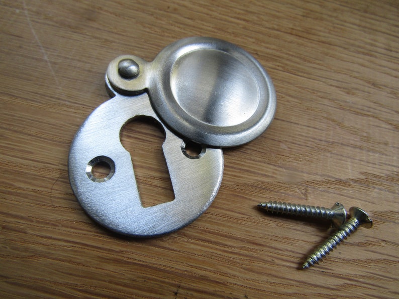 May include: A silver keyhole cover with a round, hinged lid. The cover is attached to a wooden surface with two screws.