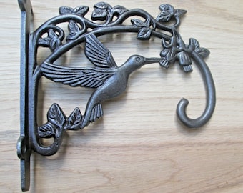 May include: Cast iron hummingbird plant hanger with a decorative floral design. The hanger features a hummingbird in flight, perched on a branch with flowers. The hanger is painted in a dark gray finish.