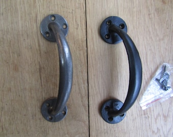 May include: Two black metal door handles with rounded edges and a curved design. The handles are mounted on a wooden surface.