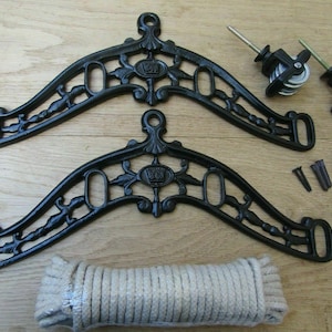 May include: A black cast iron clothesline pulley system kit. The kit includes two decorative brackets, pulleys, screws, wall anchors, and a coil of rope. The brackets have an ornate design with floral and crown details.