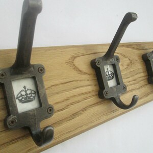 May include: A wooden coat rack with three metal hooks. Each hook has a square frame with a crown design. The hooks are mounted on a light brown, wooden plank, perfect for a hallway or entryway.