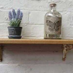 May include: A wooden shelf with gold metal brackets. The shelf is decorated with a potted lavender plant and a clear glass bottle with a butterfly design and the text "Grande Naturelle, Convallaria Majalis".