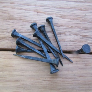 May include: A group of eight black metal nails with square heads, lying on a wooden surface. One nail is standing upright.