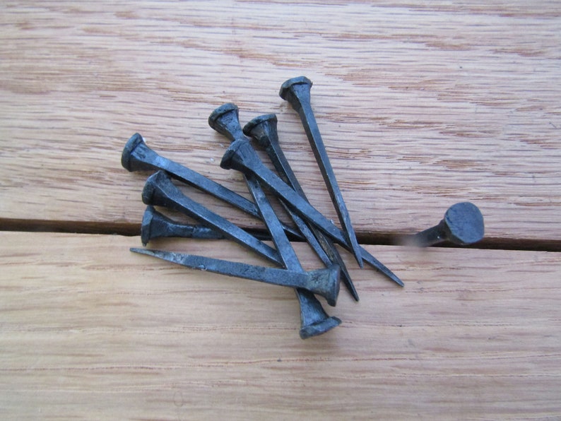 May include: A group of eight black metal nails with square heads, lying on a wooden surface. One nail is standing upright.
